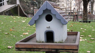 Weies Vogelhaus auf einer Wiese im Vordergrund, Kinderspielplatz im Bildhintergrund