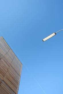 Blick nach oben auf blauen Himmel, links Ausschnitt einer Holzwand, rechts Untersicht auf lngliche Straenlampe.