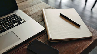Auf einem Tisch steht ein Laptop, daneben liegen ein Smartphone und ein Heft mit Stift